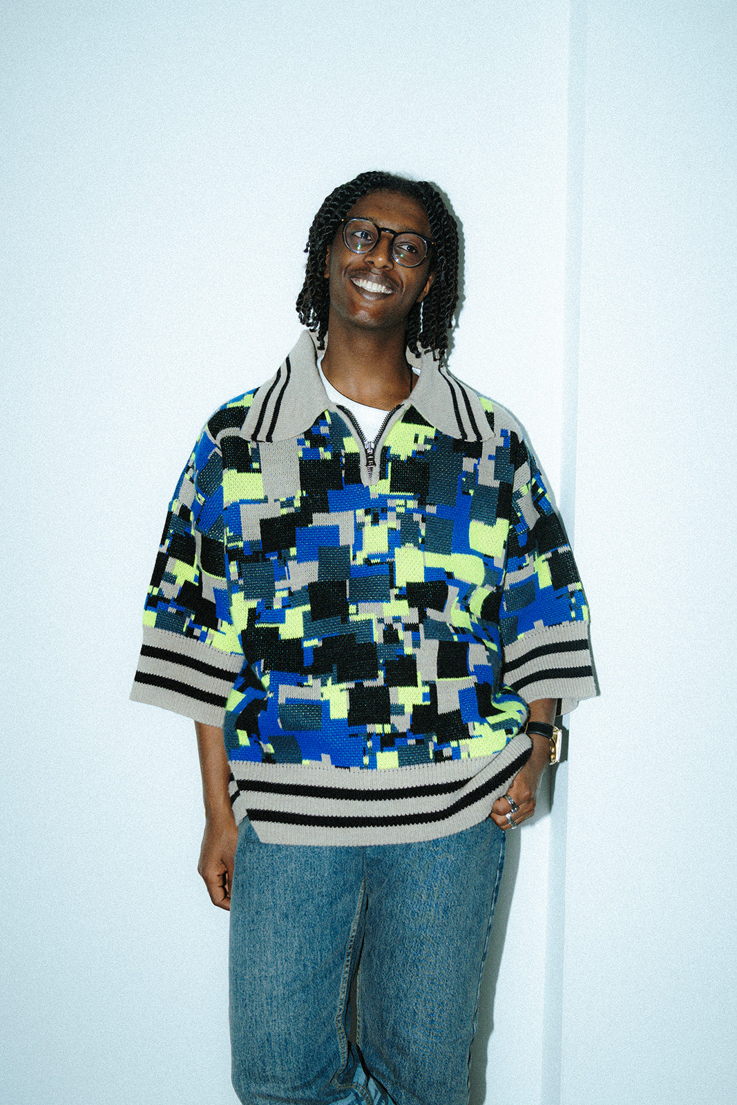 Front view of a male model wearing the GLIX "No Knit" oversized short-sleeve jacquard knit polo sweater in cobalt blue, neon yellow-green, black, and cream pixelated glitch pattern, with cream-and-black striped ribbed hem and cuffs and a cream polo collar, styled with blue jeans against a white studio background.
