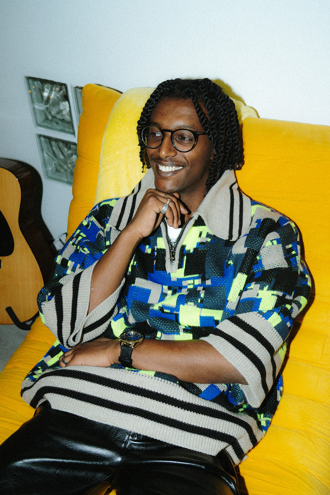 Lifestyle shot of a male model wearing the GLIX "No Knit" oversized jacquard knit polo sweater in cobalt blue, neon yellow-green, black, and cream pixelated glitch pattern, seated in a yellow armchair, showing the cream-and-black striped ribbed hem and the half-zip polo collar detail, styled with black trousers.