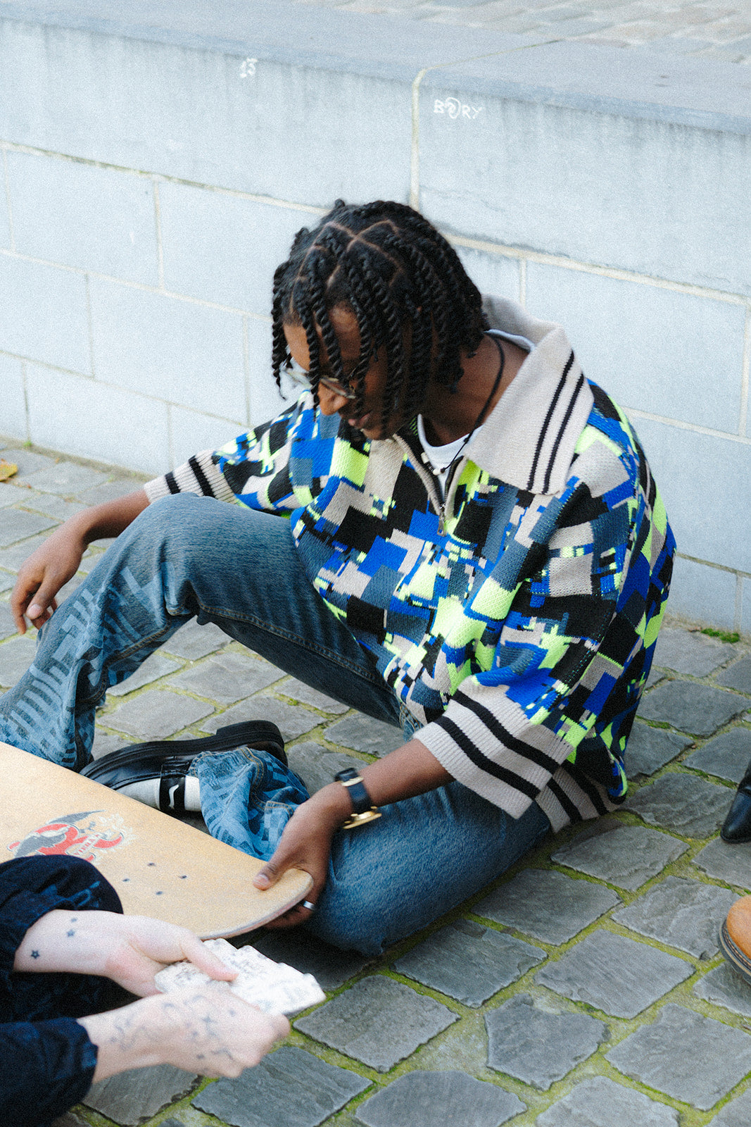 Lifestyle shot of a male model wearing the GLIX "No Knit" oversized jacquard knit polo sweater in cobalt blue, neon yellow-green, black, and cream pixelated glitch pattern, seated on cobblestone ground leaning against a wall, styled with blue jeans and black boots, holding a skateboard deck.