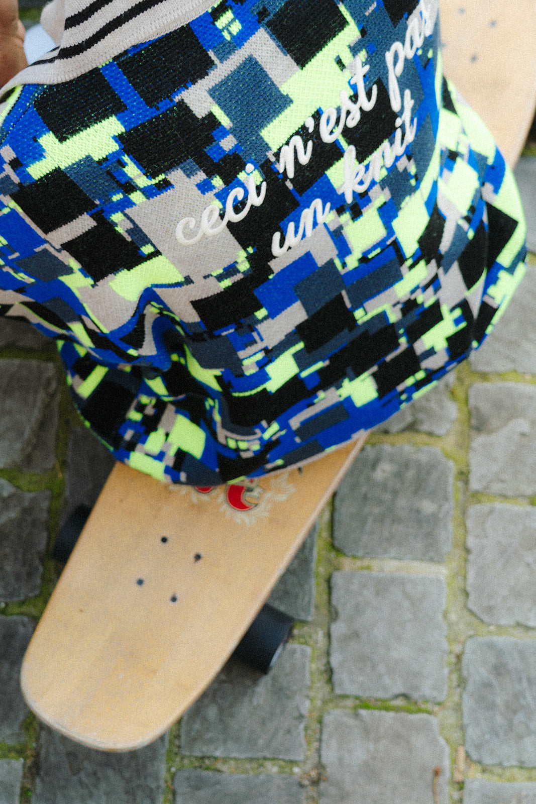 Close-up detail of the back of the GLIX "No Knit" oversized jacquard knit polo sweater, showing the all-over cobalt blue, neon yellow-green, black, and cream pixelated glitch pattern alongside the embroidered text "ceci n'est pas un knit" in white script, with a skateboard deck visible below on cobblestone ground.