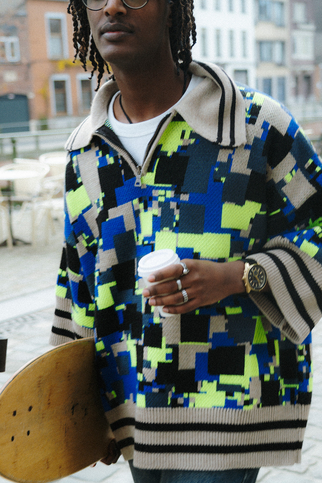 Upper-body close-up of a male model wearing the GLIX "No Knit" oversized jacquard knit polo sweater in cobalt blue, neon yellow-green, black, and cream pixelated glitch pattern, highlighting the half-zip closure, cream-and-black striped ribbed cuffs, and the volume of the oversized fit, holding a coffee cup and a skateboard on a city street.
