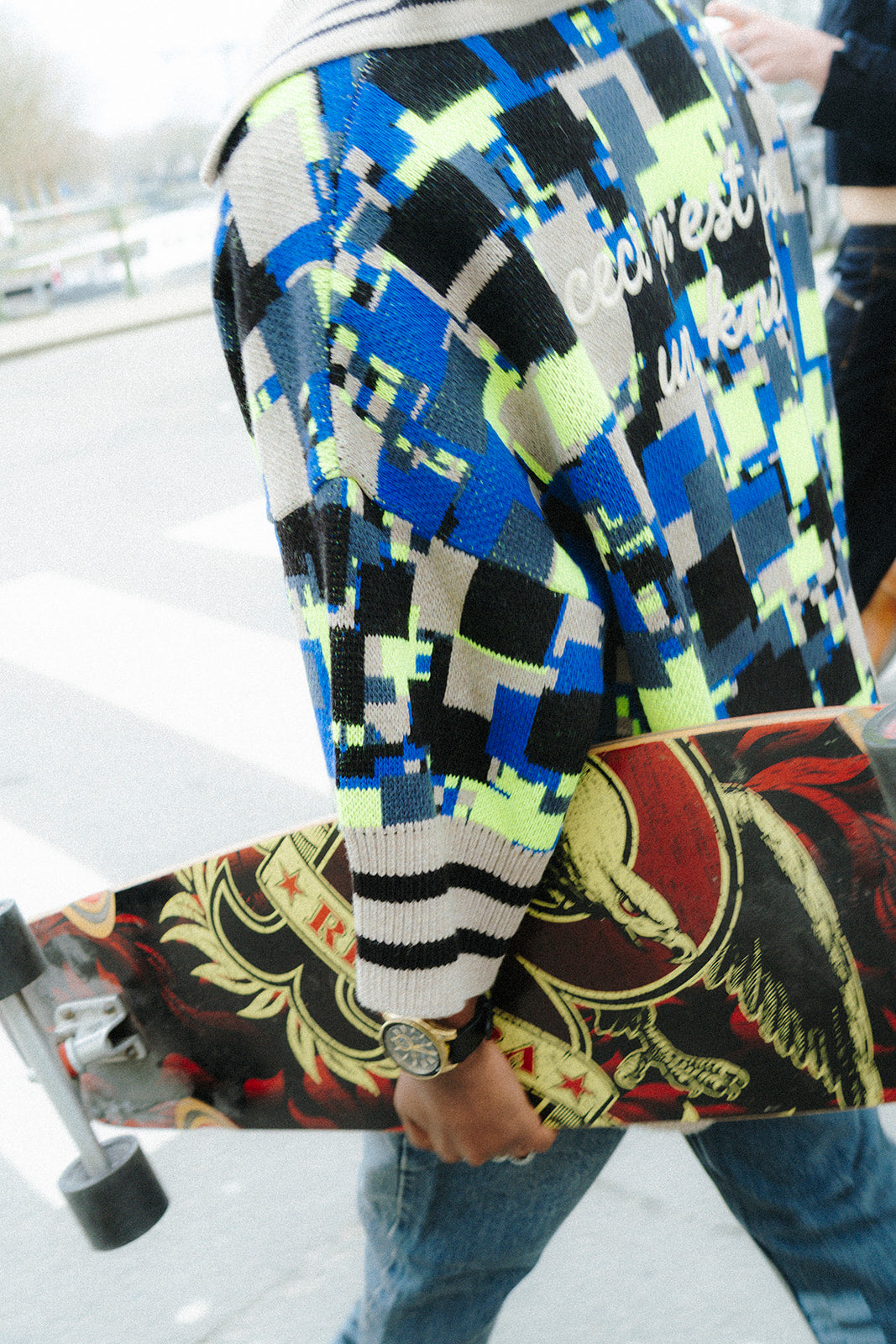 Detail shot of the sleeve and back of the GLIX "No Knit" oversized jacquard knit polo sweater in cobalt blue, neon yellow-green, black, and cream pixelated glitch pattern, showing the cream-and-black striped ribbed cuff and the embroidered "ceci n'est pas un knit" text in white script, with a decorated skateboard deck held under the arm.