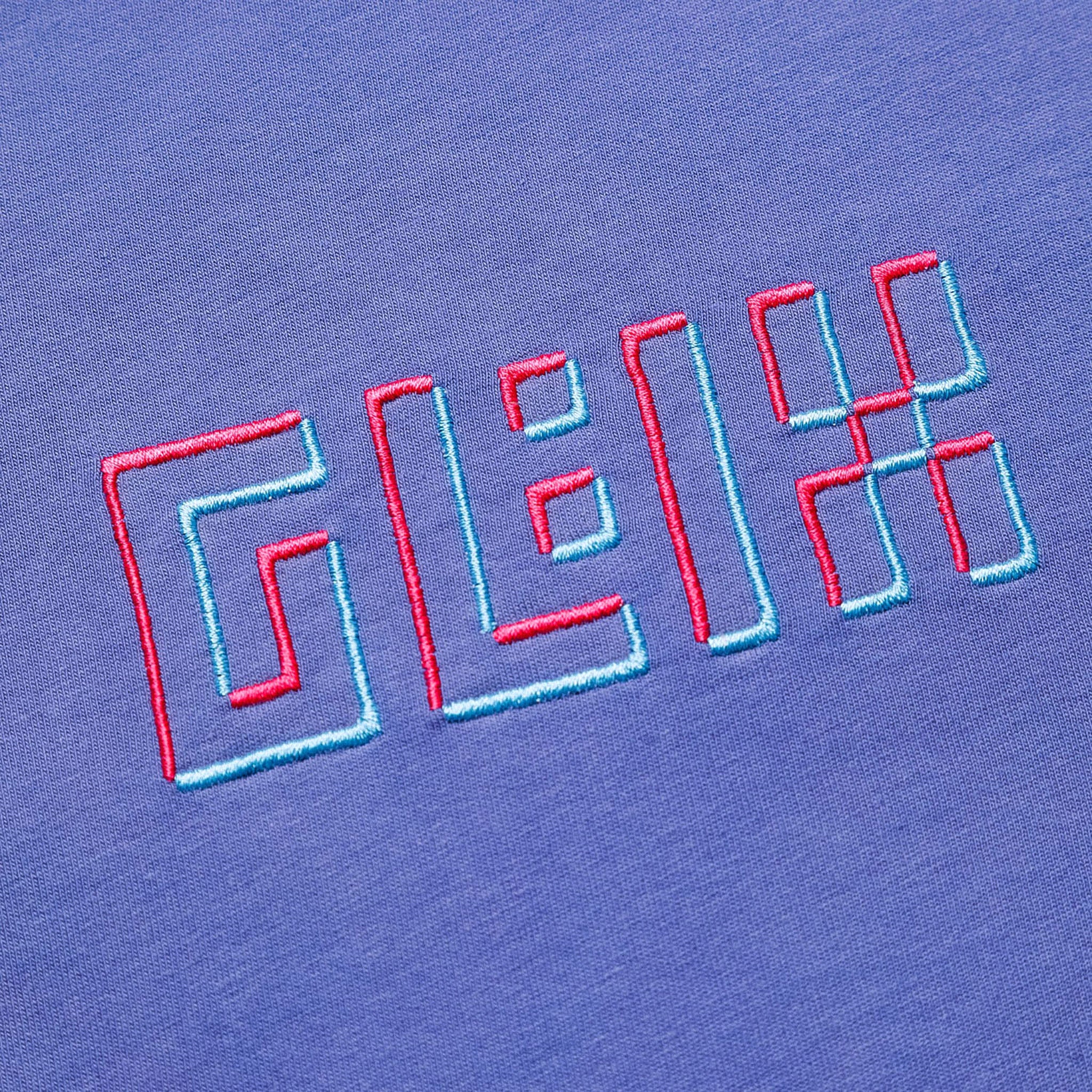 Close-up of the GLIX logo embroidered with a 3D effect in pink and light blue thread on the front of a purple oversized t-shirt.