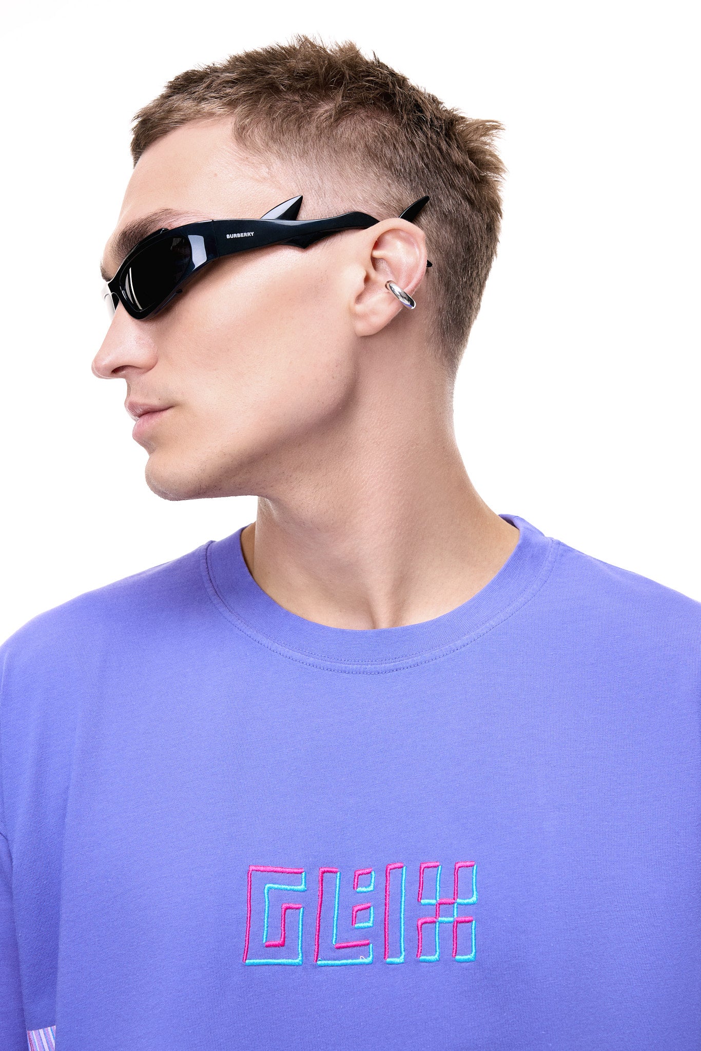 Side profile of a male model wearing the purple GLIX oversized t-shirt with a pink and blue 3D embroidered logo, styled with sharp black sunglasses and a silver ear cuff.