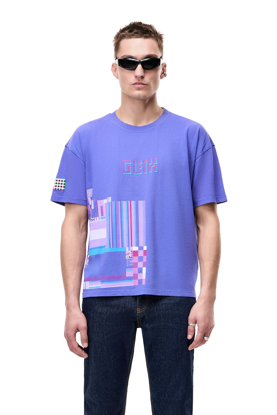 Front view of a male model wearing the purple GLIX oversized t-shirt, featuring a pink and blue 3D embroidered logo and a large geometric hem graphic, styled with black futuristic sunglasses and dark blue jeans.