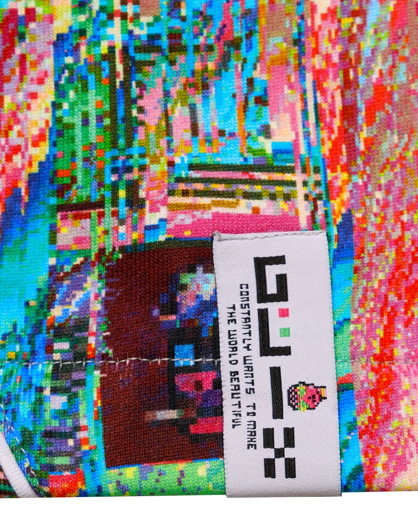 Close-up of the white woven GLIX brand tag on the "Mudderrrfall" sweatpants, featuring the brand slogan "CONSTANTLY WANTS TO MAKE THE WORLD BEAUTIFUL" against the vibrant multicolor liquid digital glitch print.