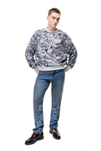 Front view of a male model wearing the GLIX "Colors' Echo" oversized sweatshirt, featuring an all-over grey and multicolor digital glitch print with ribbed grey cuffs and hem, styled with blue pixel-print denim jeans and black brogues.