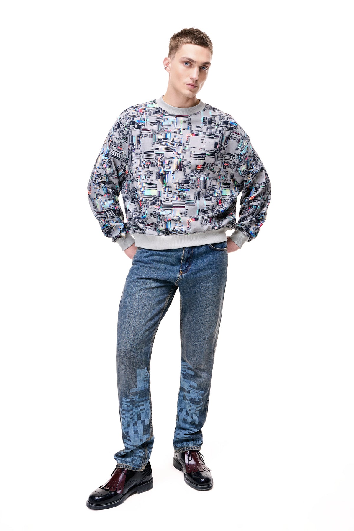 Front view of a male model wearing the GLIX "Colors' Echo" oversized sweatshirt, featuring an all-over grey and multicolor digital glitch print with ribbed grey cuffs and hem, styled with blue pixel-print denim jeans and black brogues.
