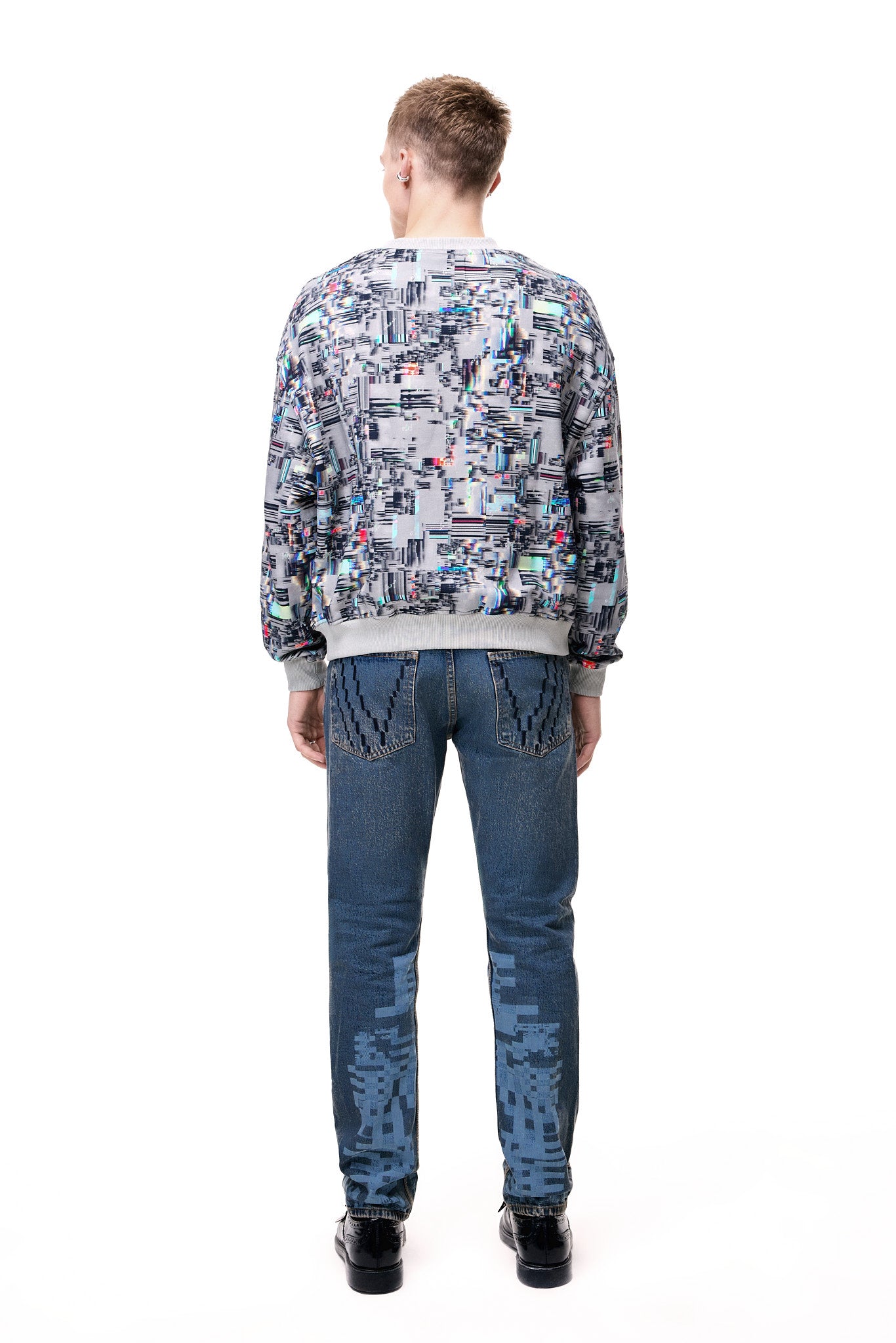 Rear view of a male model wearing the GLIX "Colors' Echo" oversized sweatshirt, showcasing the grey base with an all-over multicolor digital glitch and pixelated print, styled with blue denim jeans featuring a matching pixel pattern on the lower legs.