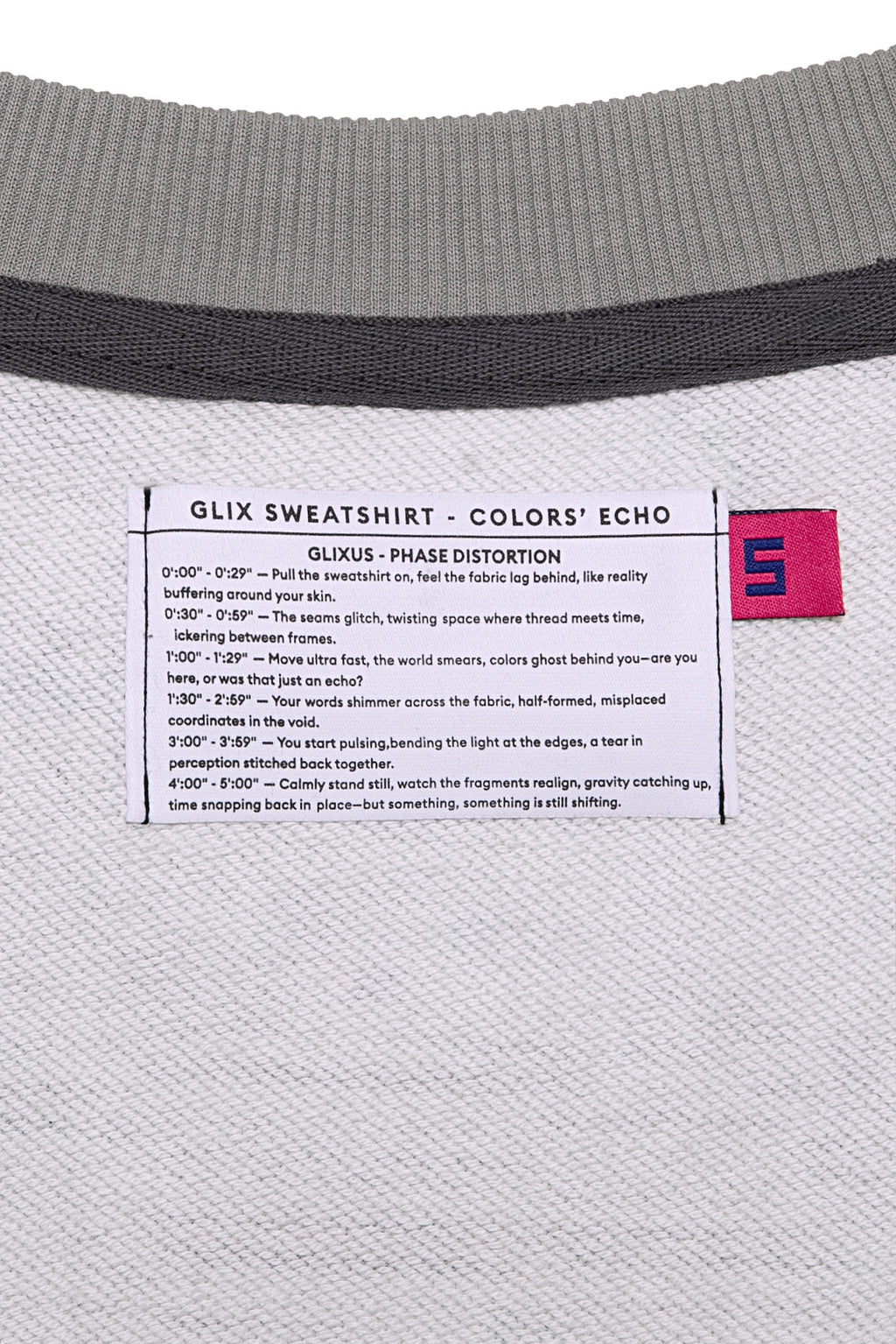 Macro close-up of the internal GLIX "Colors' Echo" white woven label featuring "GLIXUS - PHASE DISTORTION" poetic text and a pink size "S" tag, set against a grey melange sweatshirt lining.
