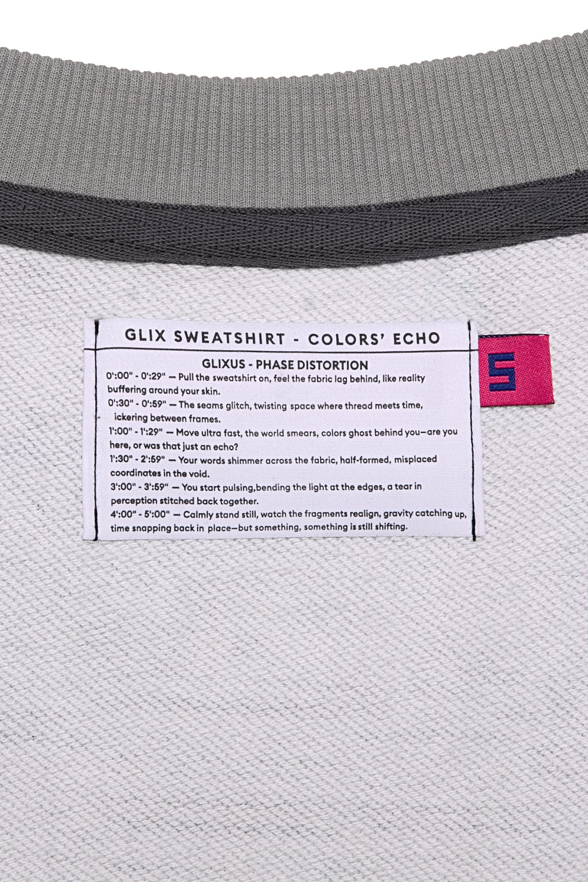 Macro close-up of the internal GLIX "Colors' Echo" white woven label featuring "GLIXUS - PHASE DISTORTION" poetic text and a pink size "S" tag, set against a grey melange sweatshirt lining.
