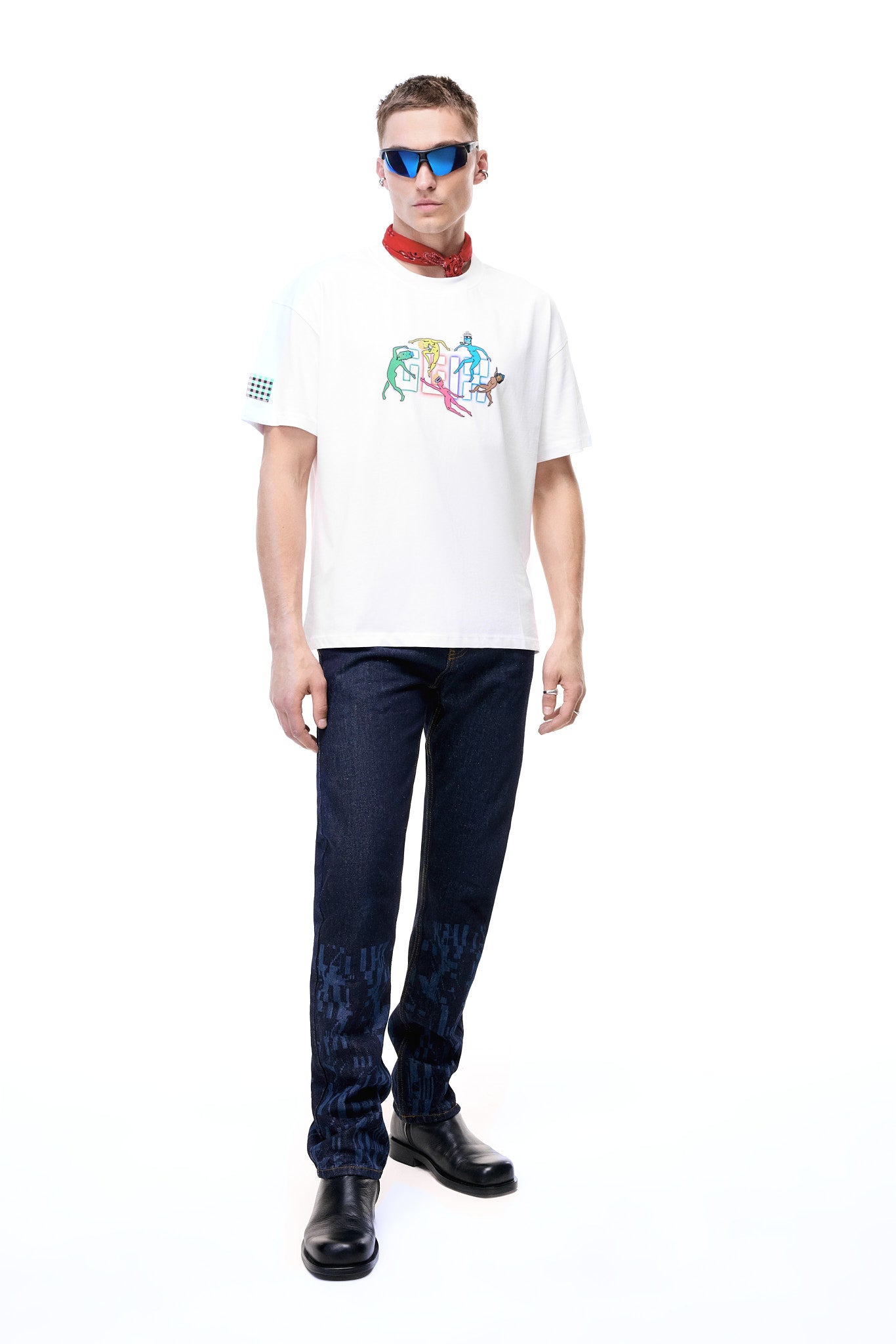 Full body shot of a male model wearing the white GLIX oversized t-shirt with a colorful "Rave" graphic, styled with dark indigo jeans, black boots, a red neck bandana, and blue mirrored sunglasses.